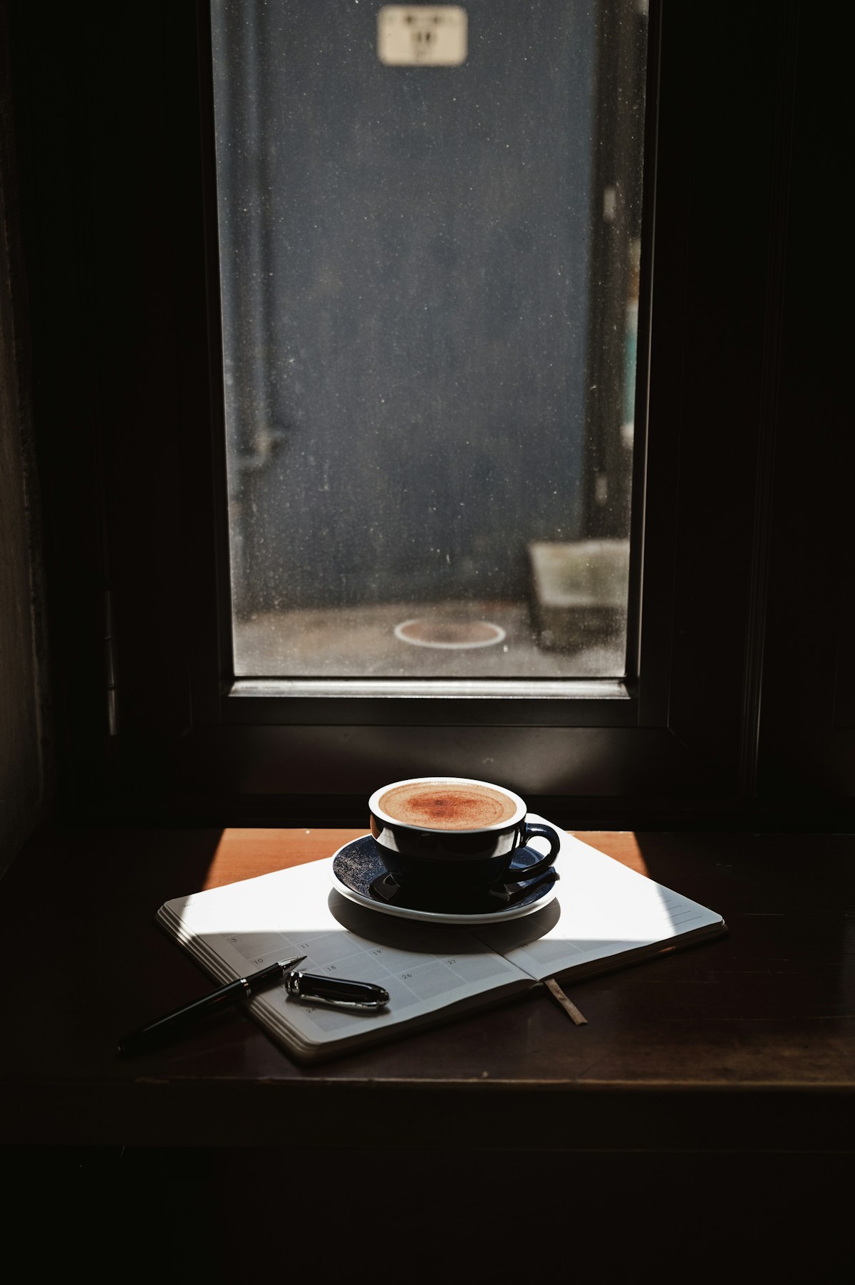 Coffee and a notebook on a wooden table beside a sunlit window